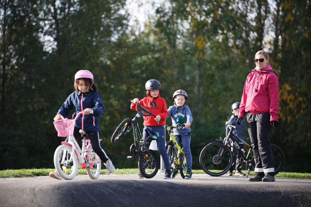 kolme pyöräilevää oppilasta pumptrack-radalla. Opettaja seuraa sivussa.
