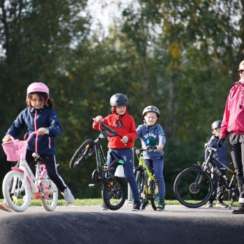 kolme pyöräilevää oppilasta pumptrack-radalla. Opettaja seuraa sivussa.